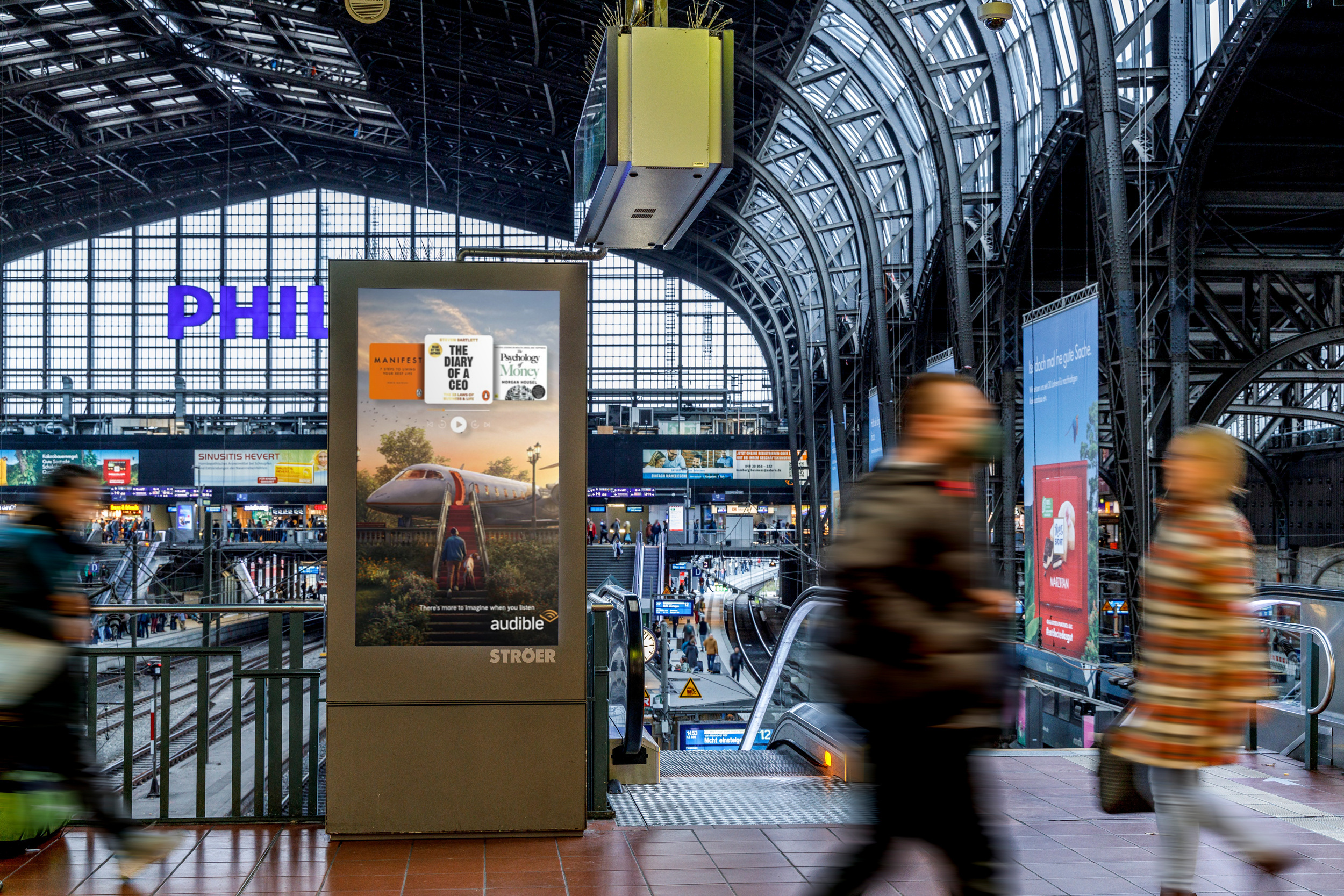 Digitale Ströer-Werbefläche für Audible mit Hörbuchcovern an einem stark frequentierten Bahnhof.