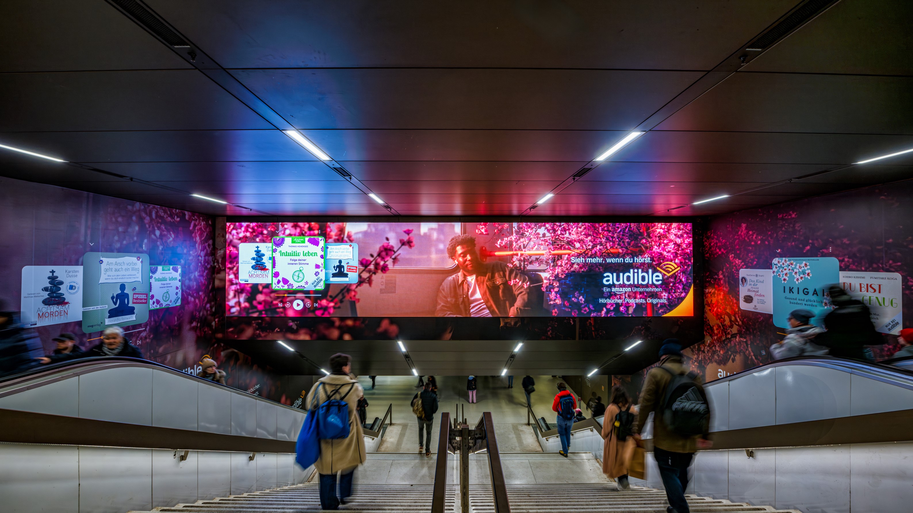 Großflächige Audible-Kampagne mit Hörbuchmotiven auf leuchtender Tunnel-Werbefläche in einem U-Bahn-Zugang.