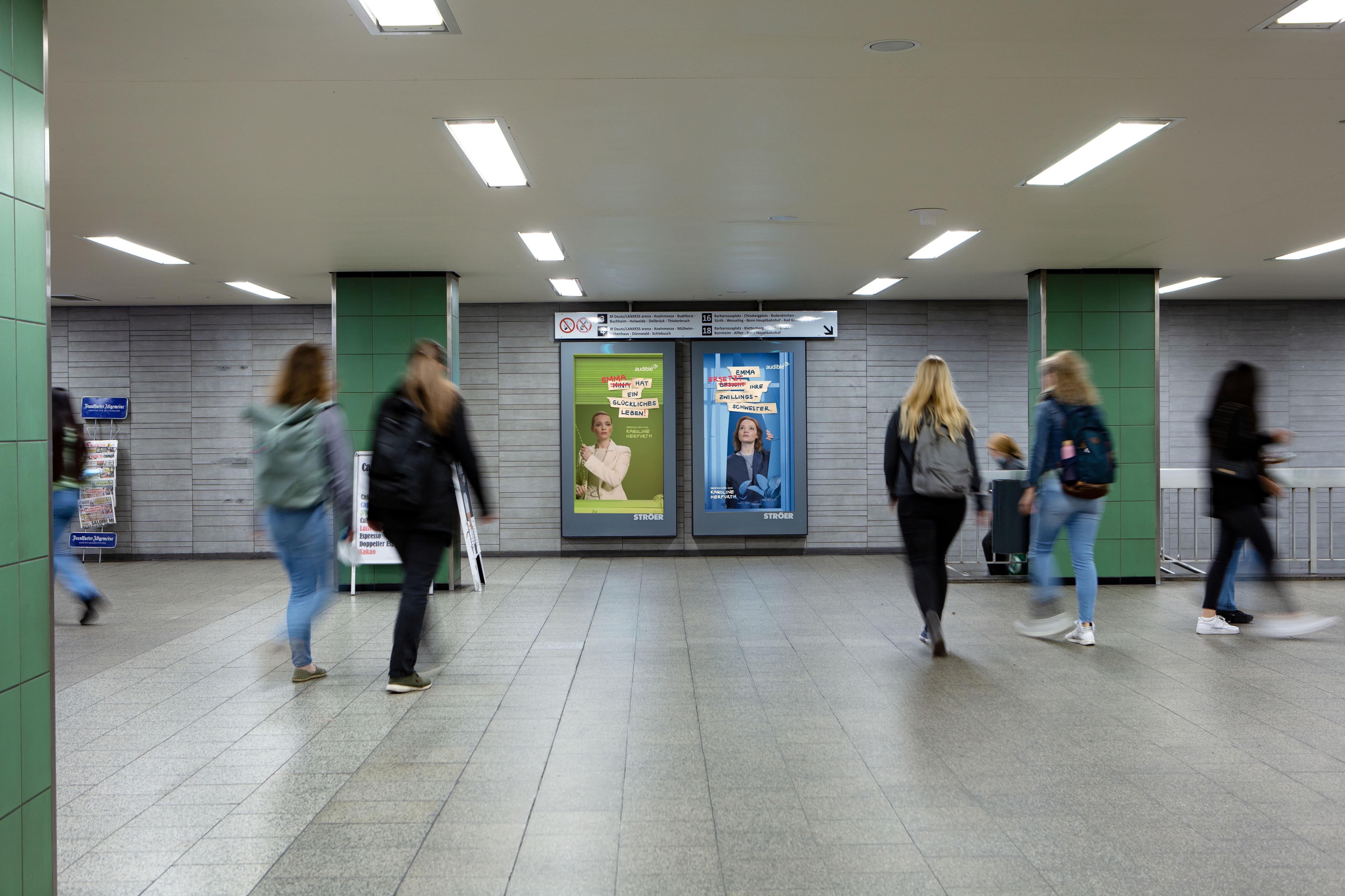Zwei digitale Ströer-Public-Video-Displays in einer U-Bahn-Station zeigen eine Werbekampagne für ein Audible-Hörbuch mit Karoline Herfurth, während verschwommene Passanten vorbeigehen.