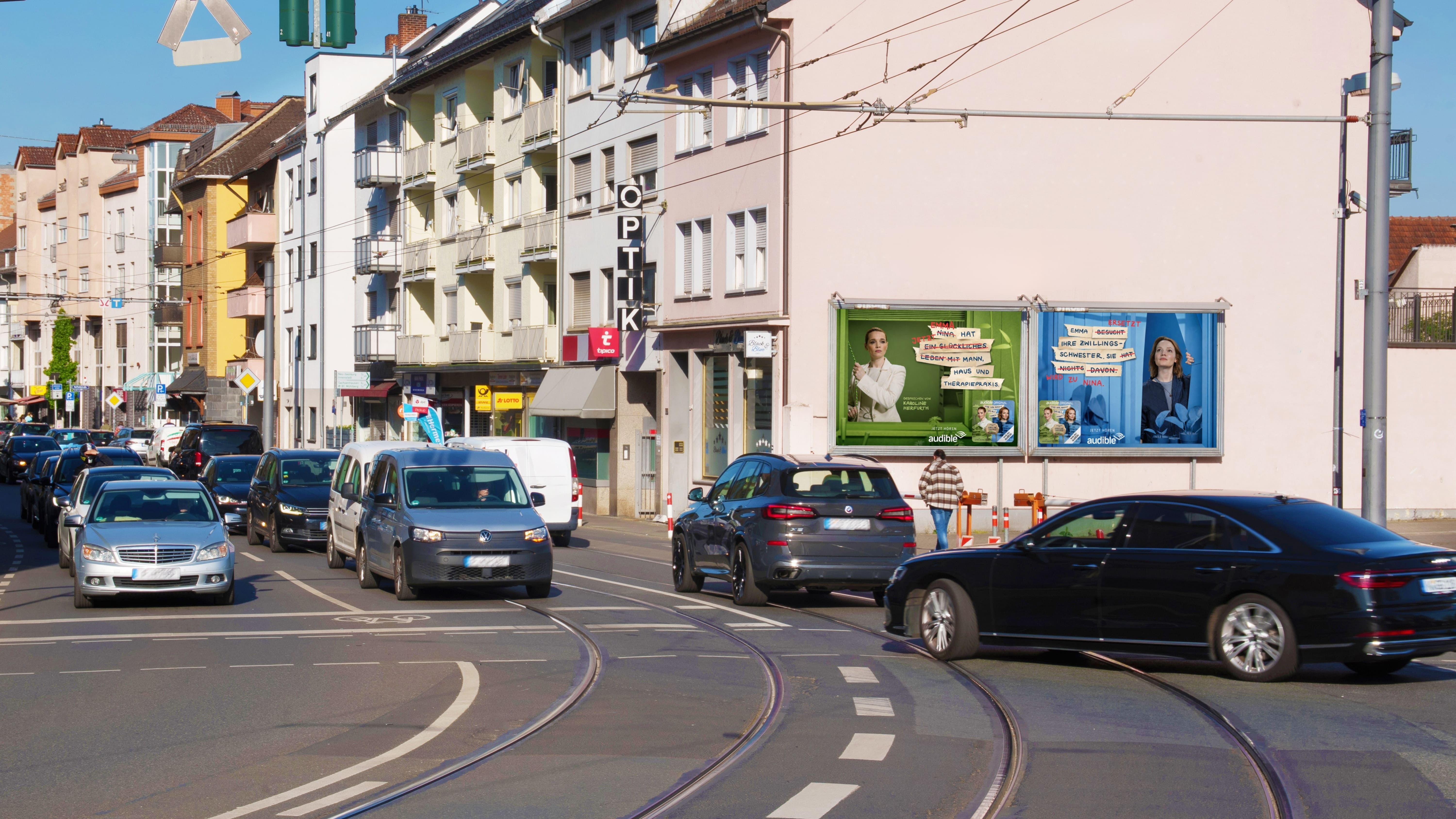Großflächige Doppel-Plakatwand an einer Hauswand an einer Straßenkreuzung mit Gleisen zeigt zwei Audible-Werbemotive.