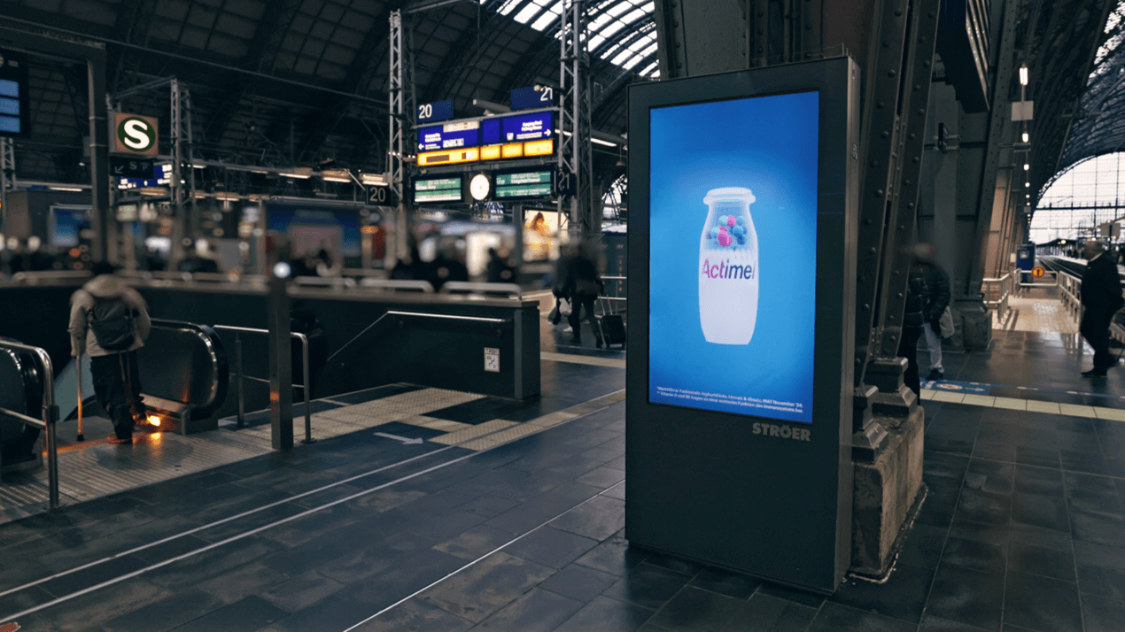 PV Station für Actimel am Bahnsteig mit Fokus auf farbigen Joghurtkulturen und Vitaminen, umgeben von Reisenden
