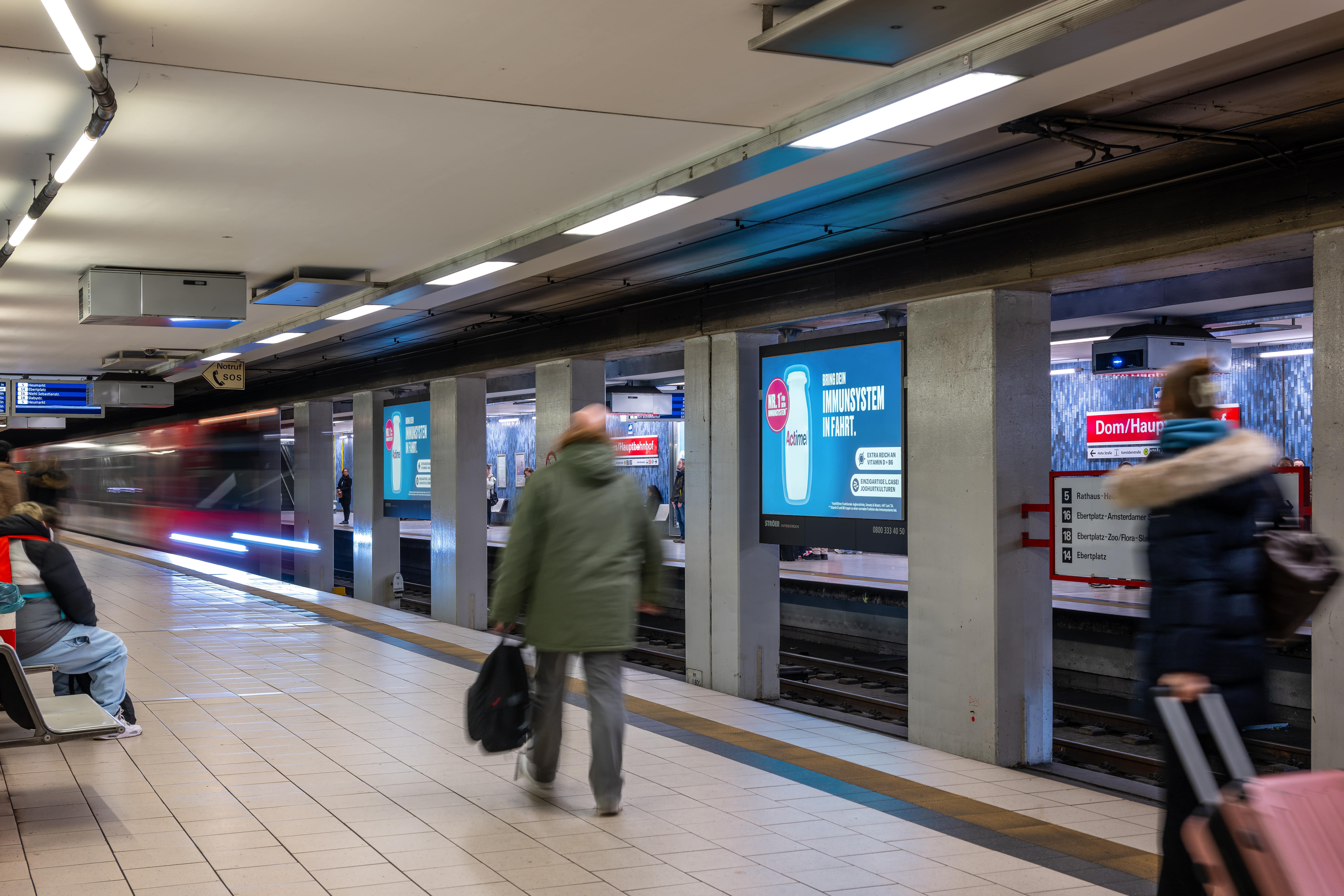 U-Bahn-Station mit Actimel-Kampagne „Bring dein Immunsystem in Fahrt“ auf einem Infoscreen, sichtbar für wartende Fahrgäste auf beiden Bahnsteigseiten