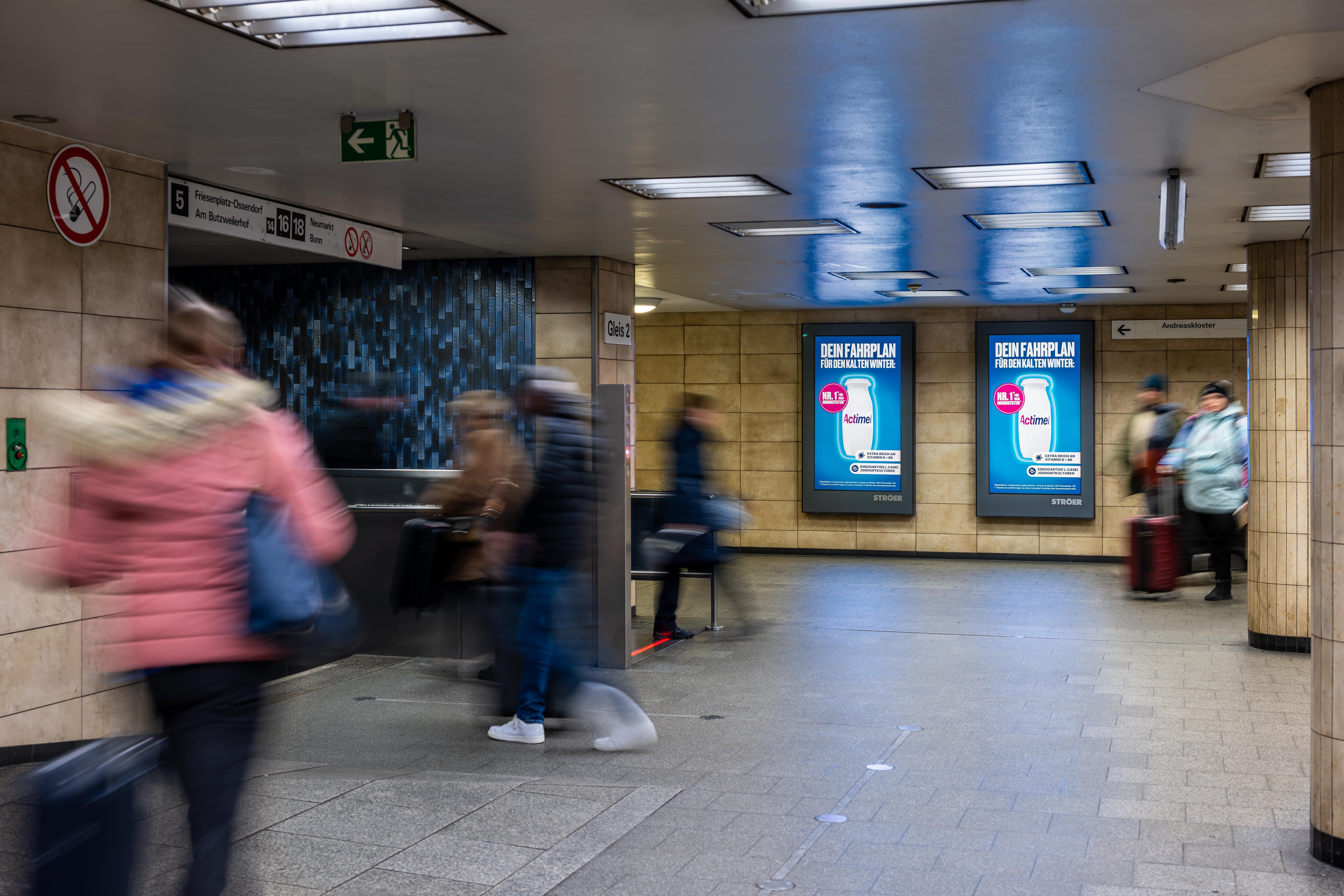 Actimel-Werbung in einer Unterführung mit Fokus auf Erkältungssaison, flankiert von Passant:innen mit Koffern und Winterkleidung