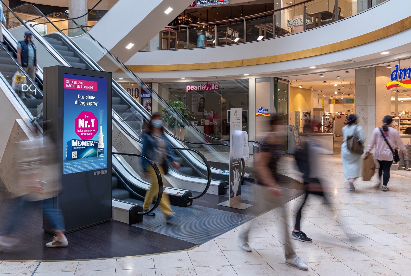 Digitale Werbeanzeige für MometaHEXAL® in Einkaufszentrum – In einem belebten Shoppingcenter neben einer Rolltreppe zeigt ein Bildschirm eine Anzeige für MometaHEXAL® mit der Botschaft „Das blaue Allergiespray“.