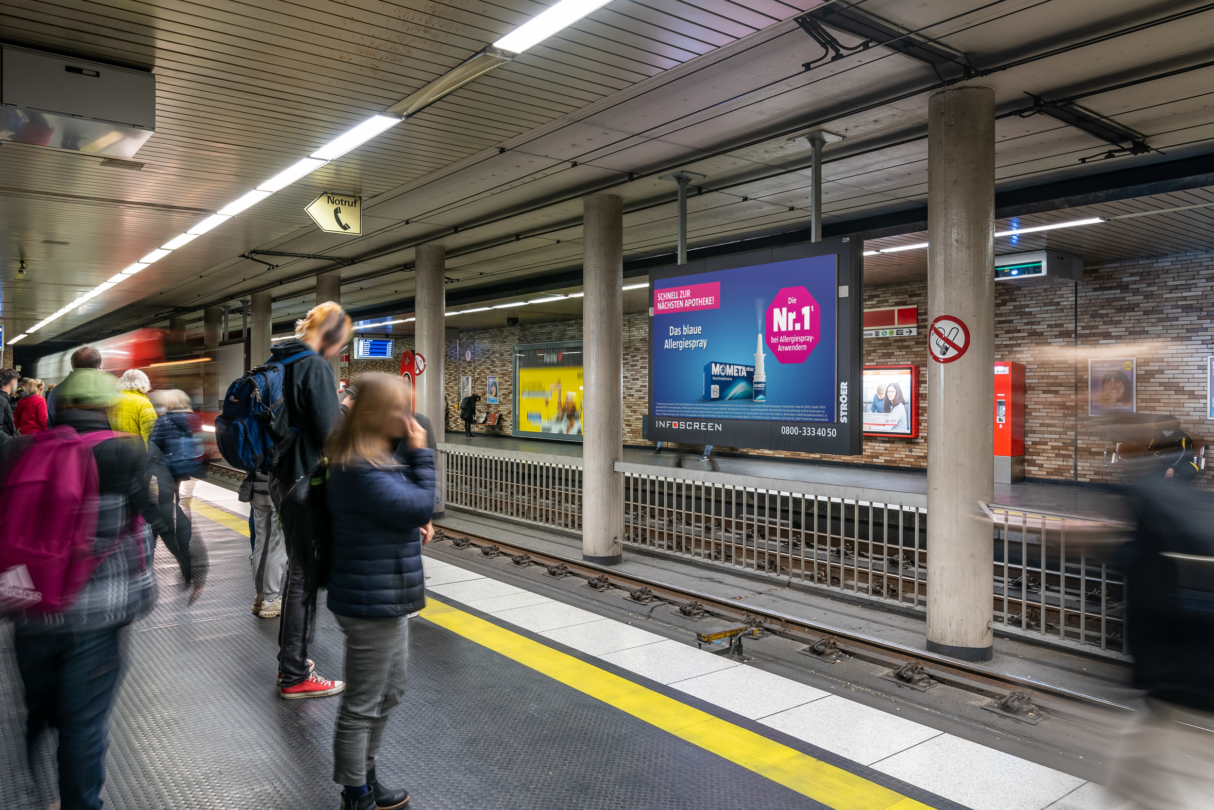 DOOH-Werbung für MometaHEXAL® in U-Bahn-Station – Fahrgäste gehen an einem digitalen Werbescreen vorbei, der das Allergiespray MometaHEXAL® mit dem Slogan „Sprühe Deinen Heuschnupfen weg!“ zeigt.