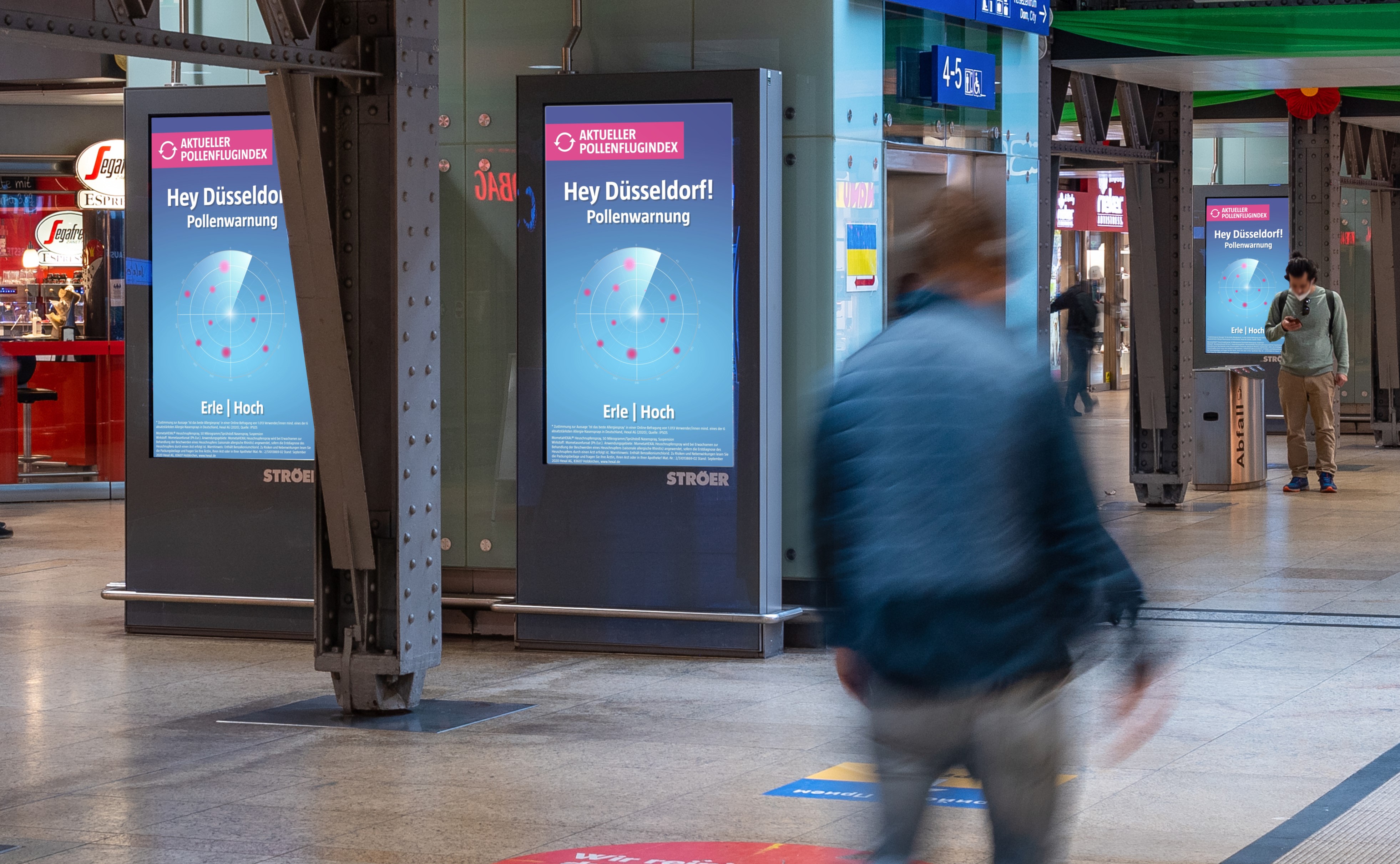 Digitale Werbedisplays in Bahnhof mit aktueller Pollenflugwarnung – Mehrere Bildschirme in einem Bahnhof zeigen eine individuelle Pollenwarnung für Düsseldorf mit der Belastung für Erle.