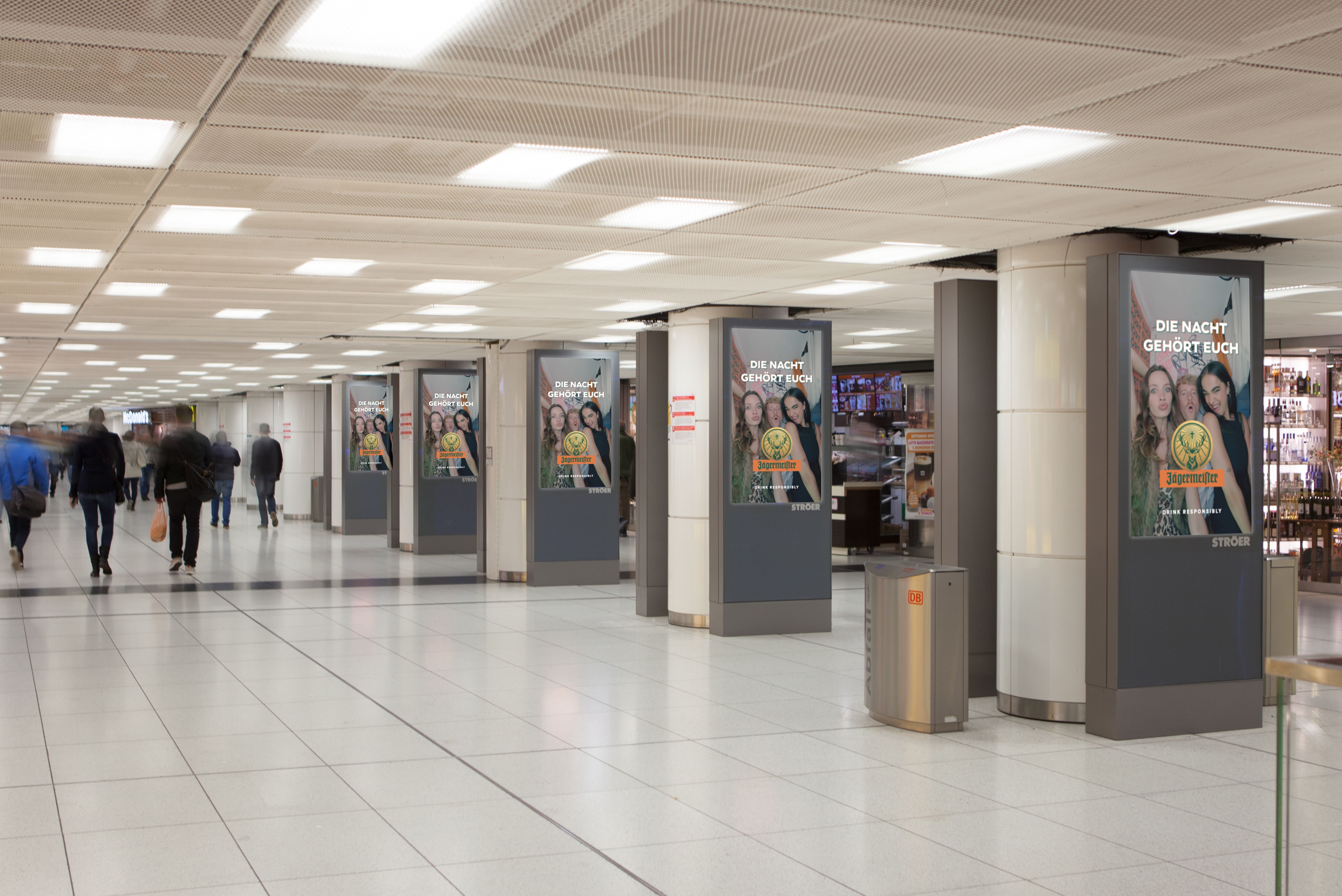 Mehrfache digitale Werbeträger mit Werbung von Jägermeister in stark frequentierter Bahnhofspassage, symmetrisch platziert für maximale Markenpräsenz im Laufweg der Pendler.