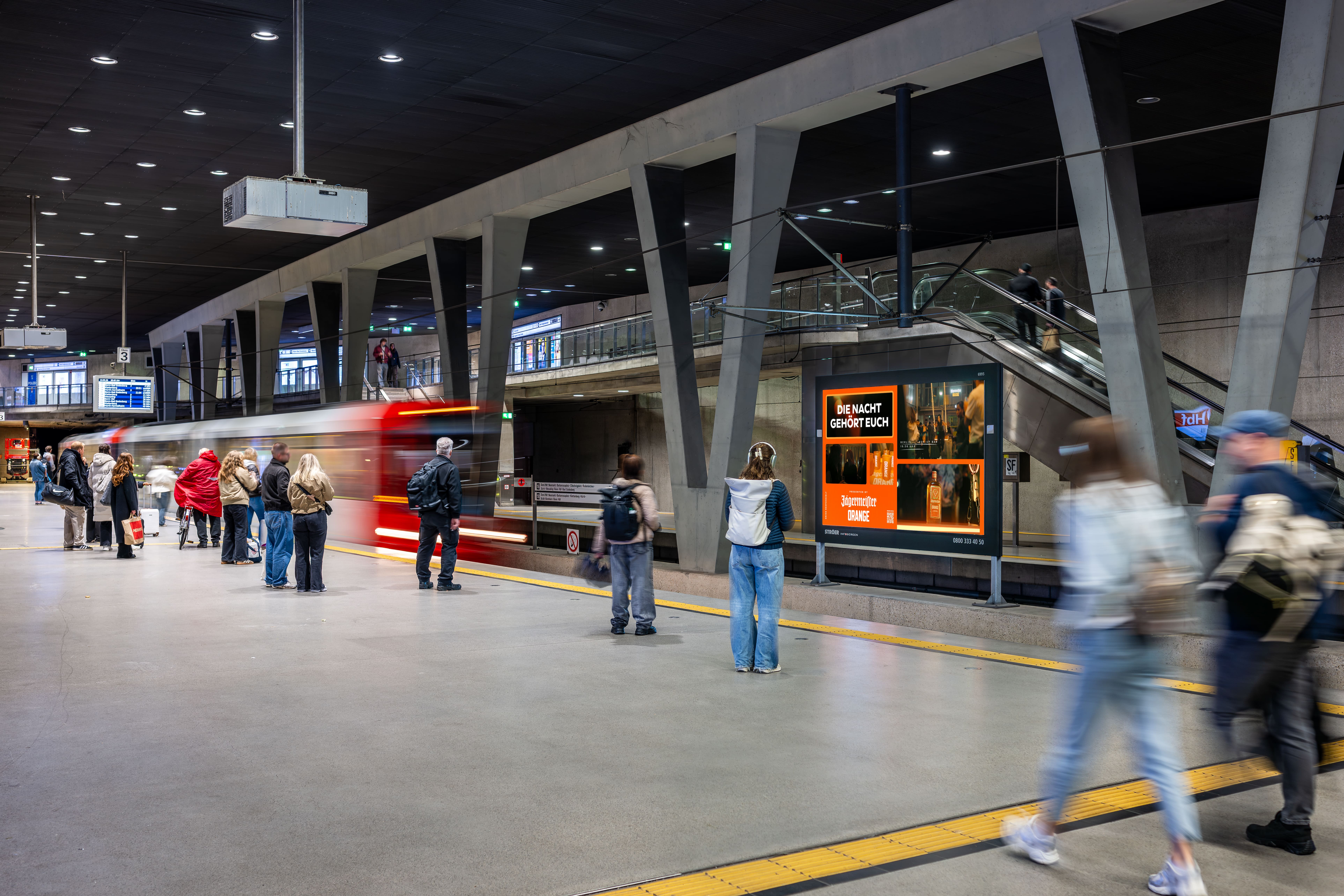 Bahnsteig einer modern gestalteten Kölner U-Bahn-Station mit grauen Betonpfeilern. Auf der linken Seite stehen wartende Fahrgäste, während eine rot-weiße U-Bahn in dynamischer Unschärfe vorbeifährt. Auf der rechten Seite steht ein großer, leuchtender digitaler Werbescreen mit einer orangefarbenen Jägermeister-Werbung und dem Slogan „Die Nacht gehört euch“. Im Vordergrund laufen Passanten vorbei.