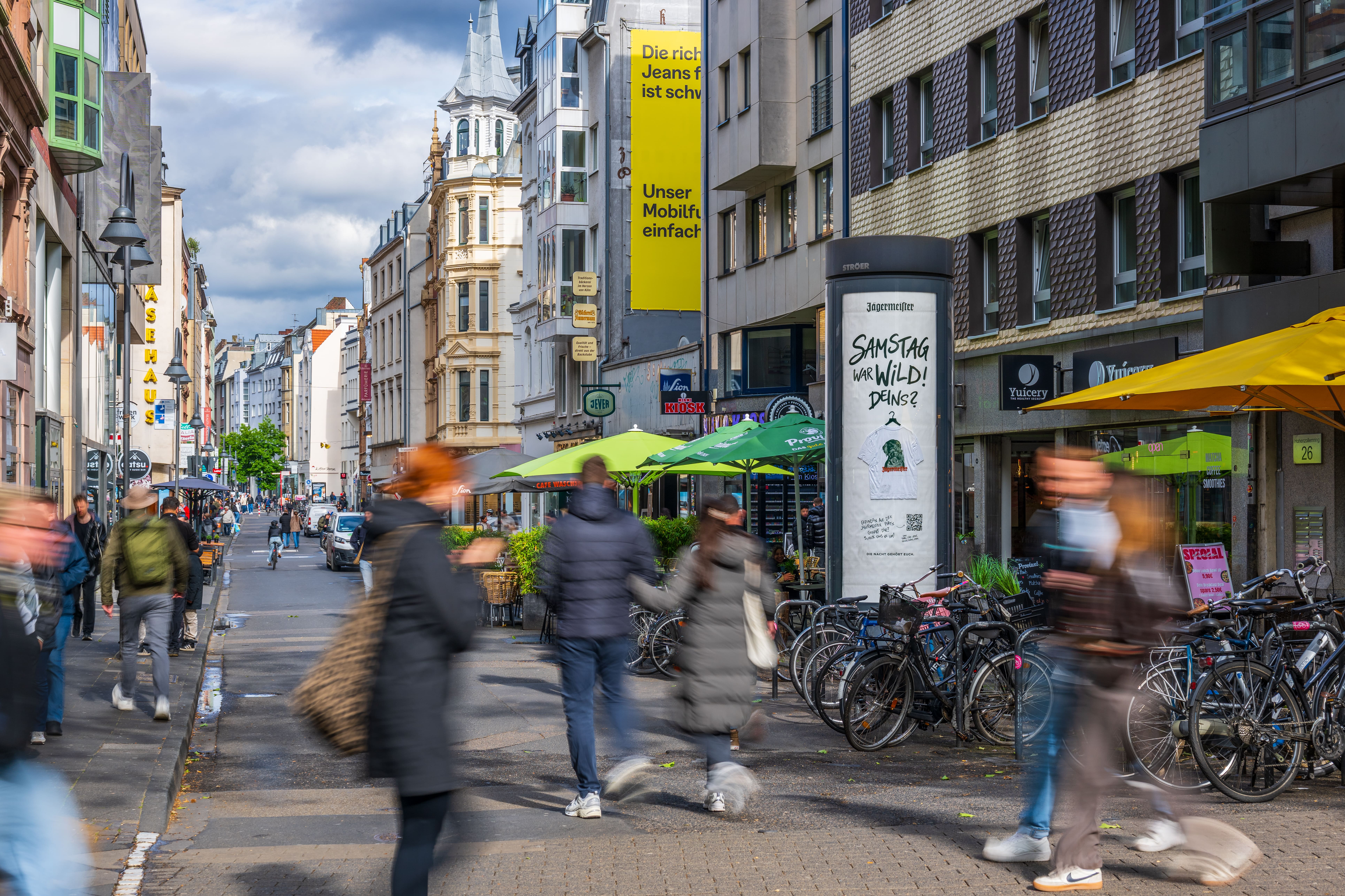 Blick in die belebte Kölner Ehrenstraße mit Fußgängern, Geschäften und Außengastronomie unter grünen Sonnenschirmen. Auf der rechten Straßenseite steht eine graue Werbesäule mit einem weißen Plakat, auf dem ein bedrucktes T-Shirt abgebildet ist. Darüber steht der handschriftliche Schriftzug „Samstag war wild! Deins?“. Die Passanten im Vordergrund sind durch Bewegung leicht verschwommen.