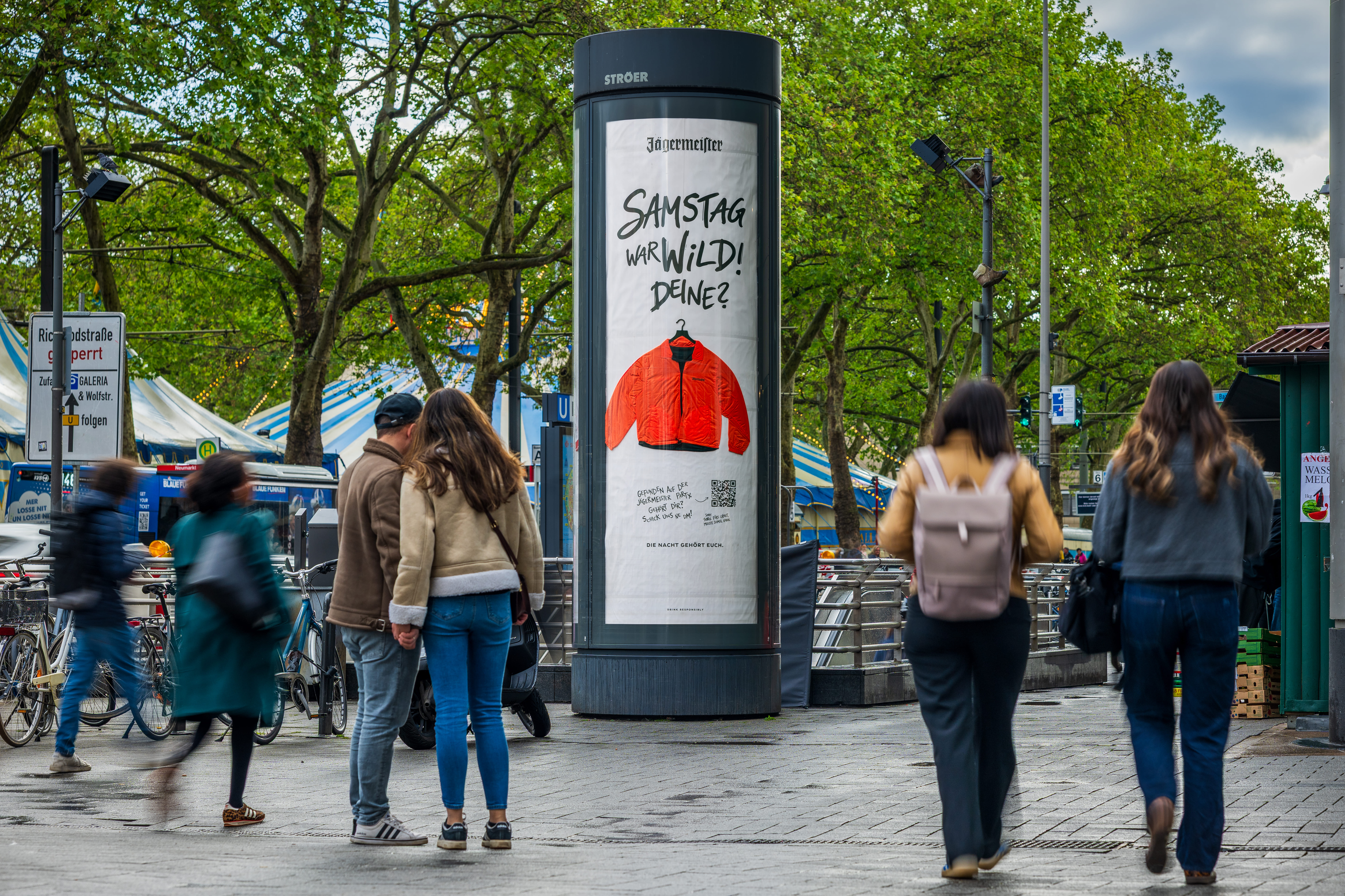 Eine große Litfaßsäule auf einem gepflasterten Platz am Kölner Neumarkt vor einer Kulisse aus grünen Bäumen. Auf der Säule klebt ein weißes Werbeplakat von Jägermeister mit einer abgebildeten orangefarbenen Winterjacke und der Frage „Samstag war wild! Deins?“. Mehrere Passanten laufen im Vordergrund an der Säule vorbei.
