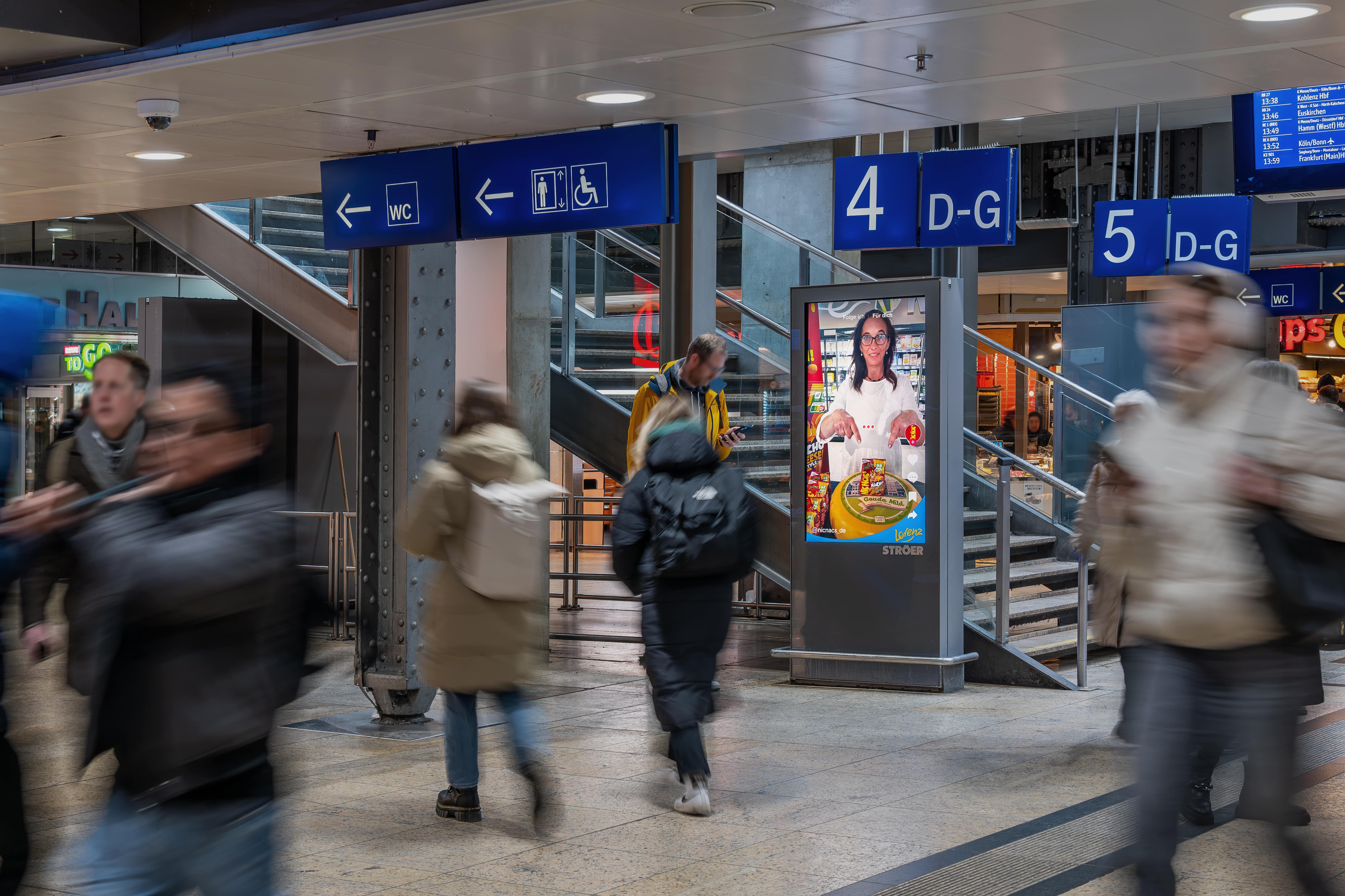 Käse-Prinzessin als TikTok-Testimonial in digitaler Bahnhofs-Werbung von Lorenz NicNac’s, platziert zwischen Gleiszugängen mit Laufpublikum