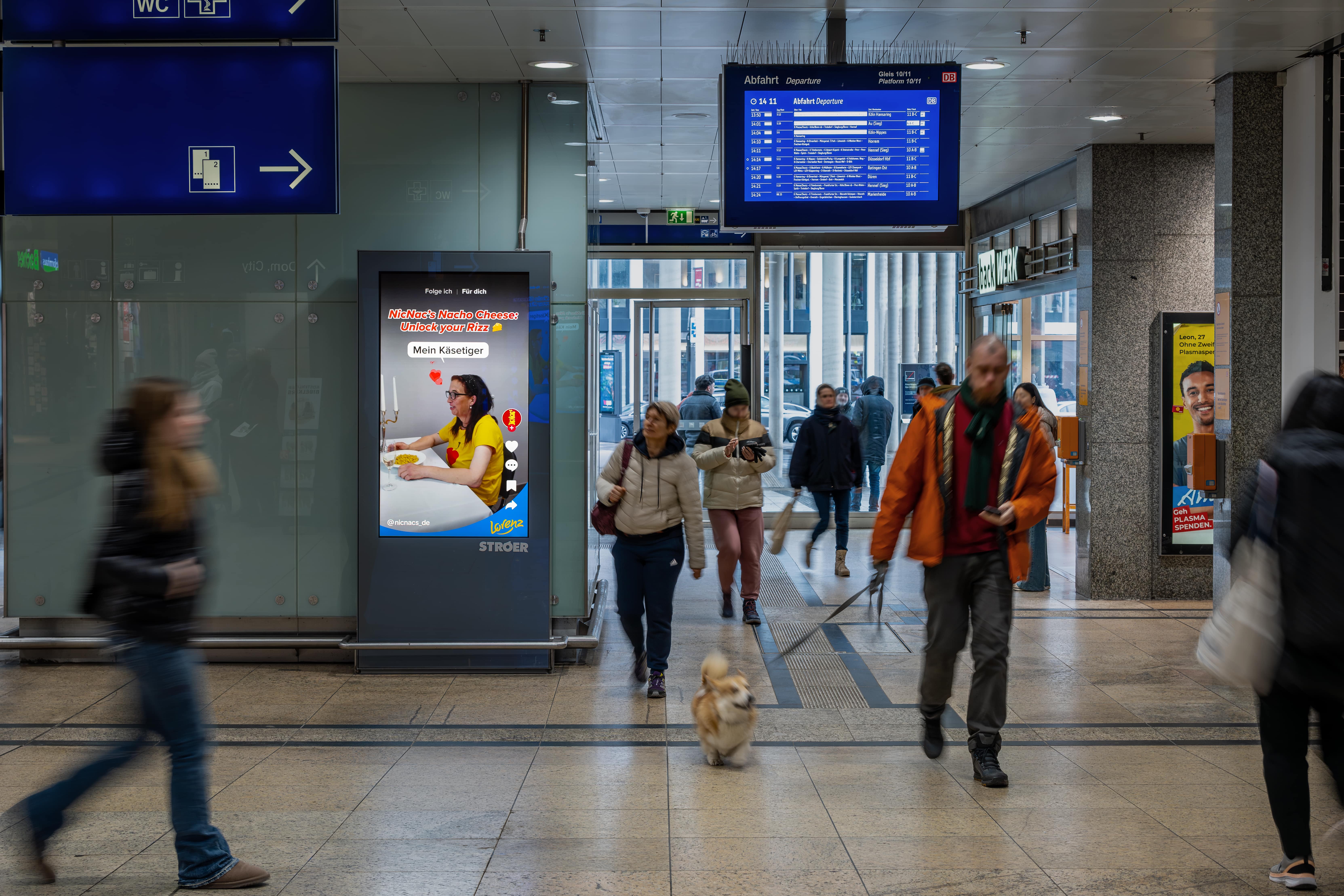 Einzelne PV Station im Bahnhof zeigt „Mein Käsetiger“-Kampagnenmotiv mit TikTok-Optik und vielen Reisenden im Bild