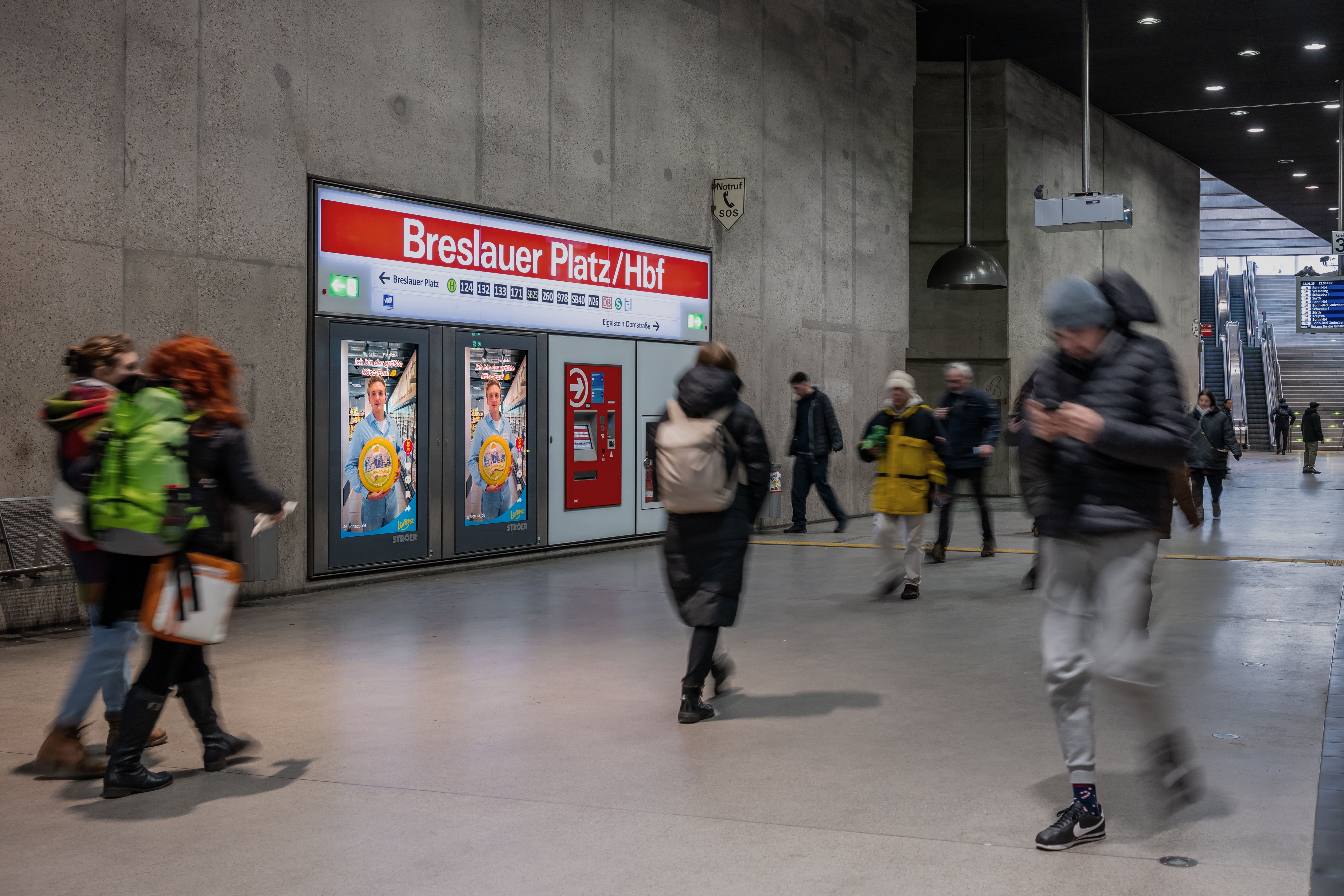 Digitale OOH-Werbung für NicNac’s Nacho Cheese Style mit Ludger Bökelmann am Breslauer Platz/Hbf, umgeben von Pendler:innen
