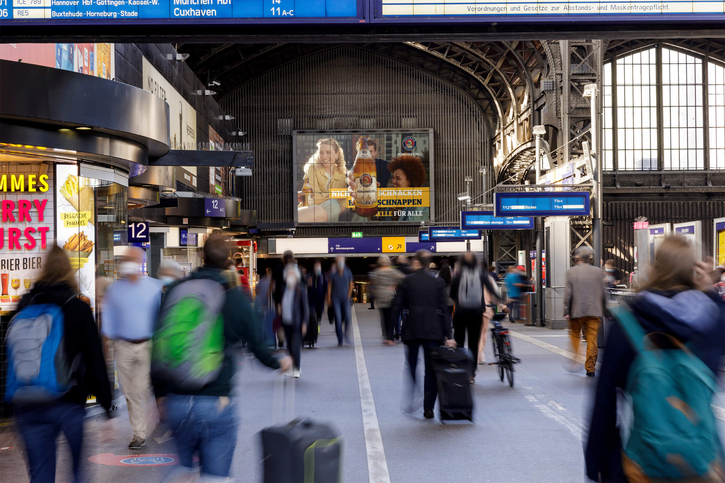 Digitale Out-of-Home-Werbung auf großformatigem Screen mit Werbung von Paulaner Münchner Hell im Durchgang des Hauptbahnhofs Hamburg, stark frequentiert von Reisenden und Pendlern.