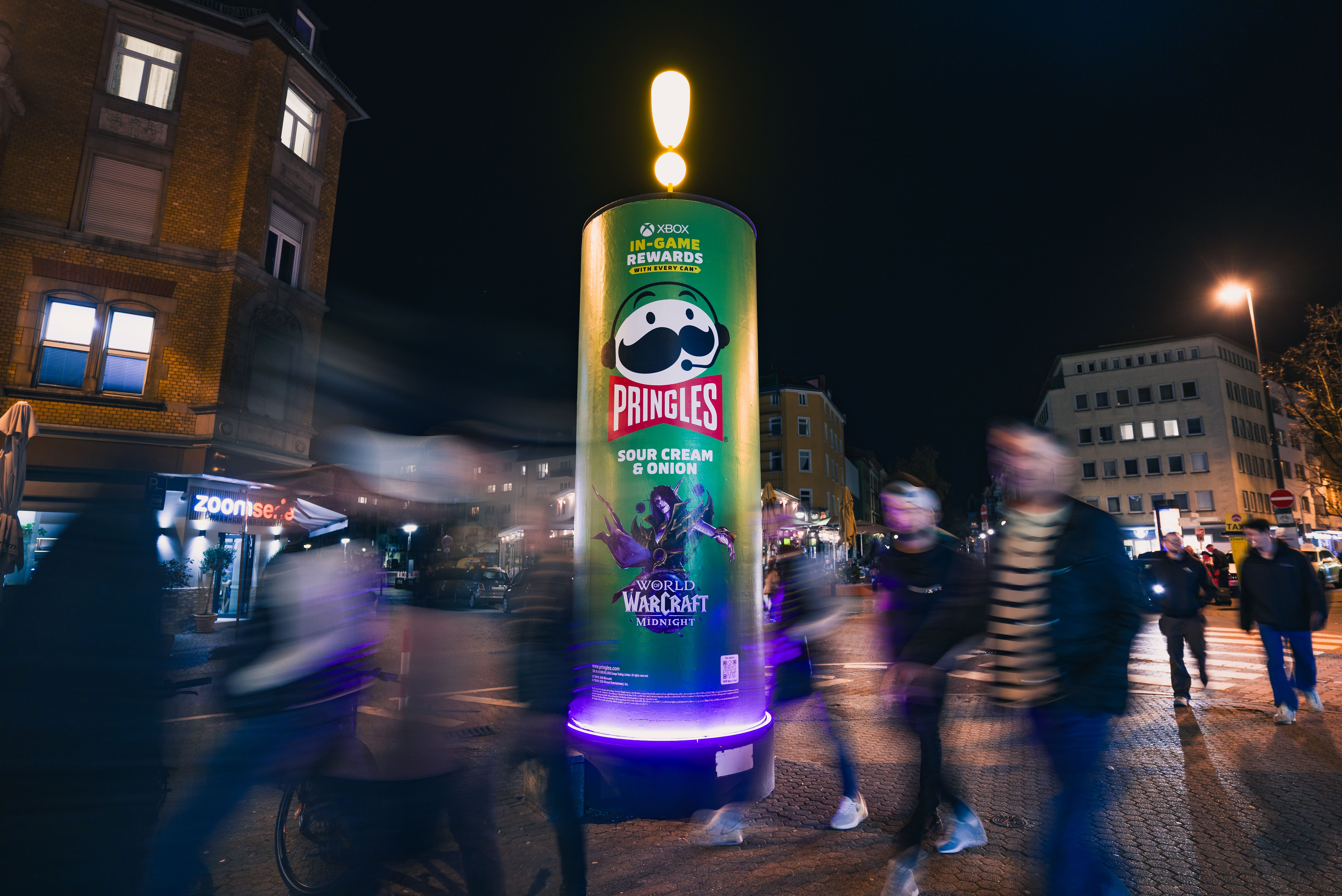 Außenaufnahme bei Dunkelheit mit Fokus auf eine grüne Pringles-Werbesäule mit leuchtender Spitze und violettem Sockellicht. Mehrere Fußgänger kreuzen das Bild im Vordergrund mit sichtbarer Bewegungsunschärfe vor urbaner Hintergrundkulisse.