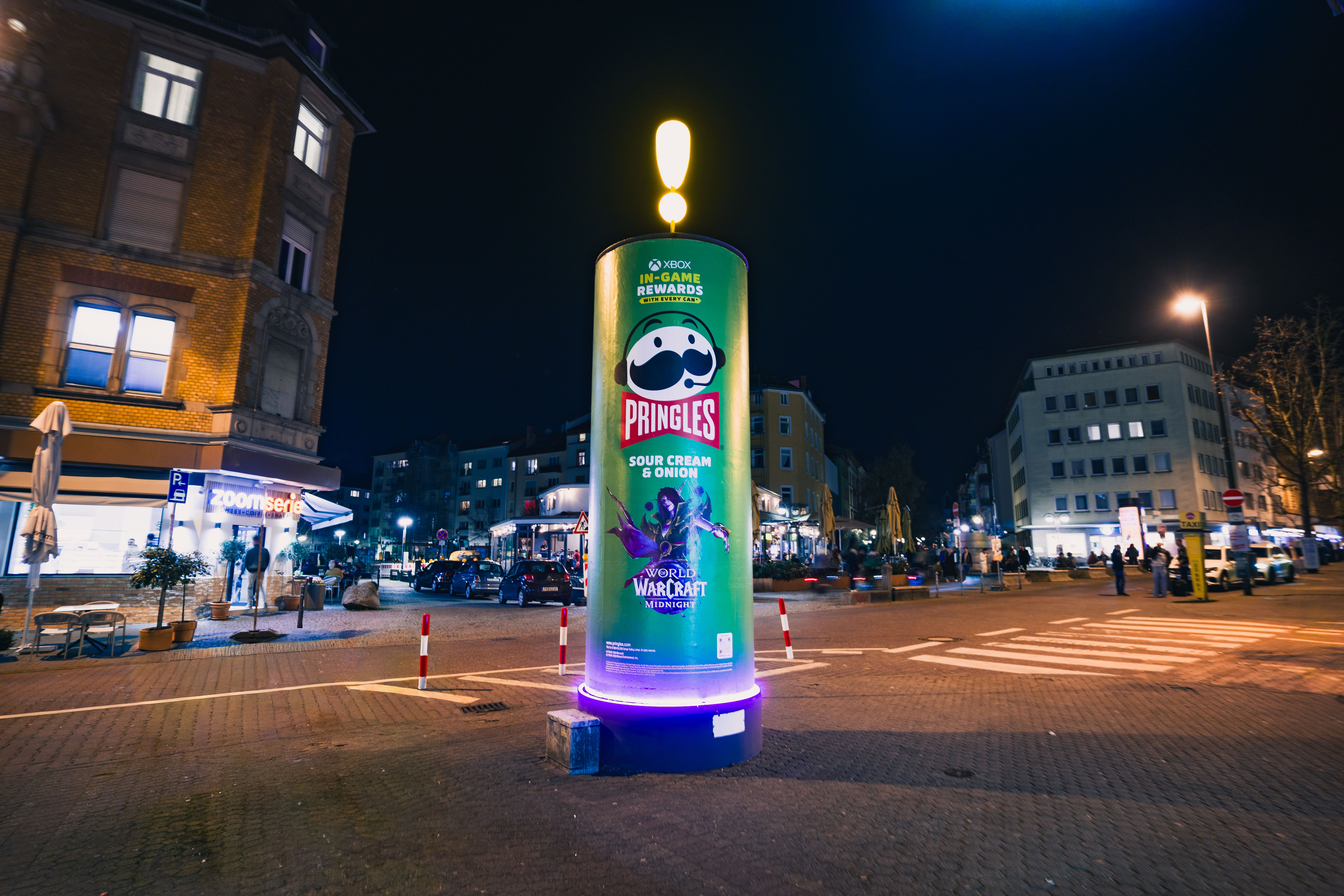 Totale einer beleuchteten Werbesäule im Design einer Pringles-Dose auf einem gepflasterten Stadtplatz bei Nacht. Die Säule mit einem strahlenden Ausrufezeichen auf der Spitze steht frei im Raum nahe einem Fußgängerüberweg und beleuchteter Gastronomie.