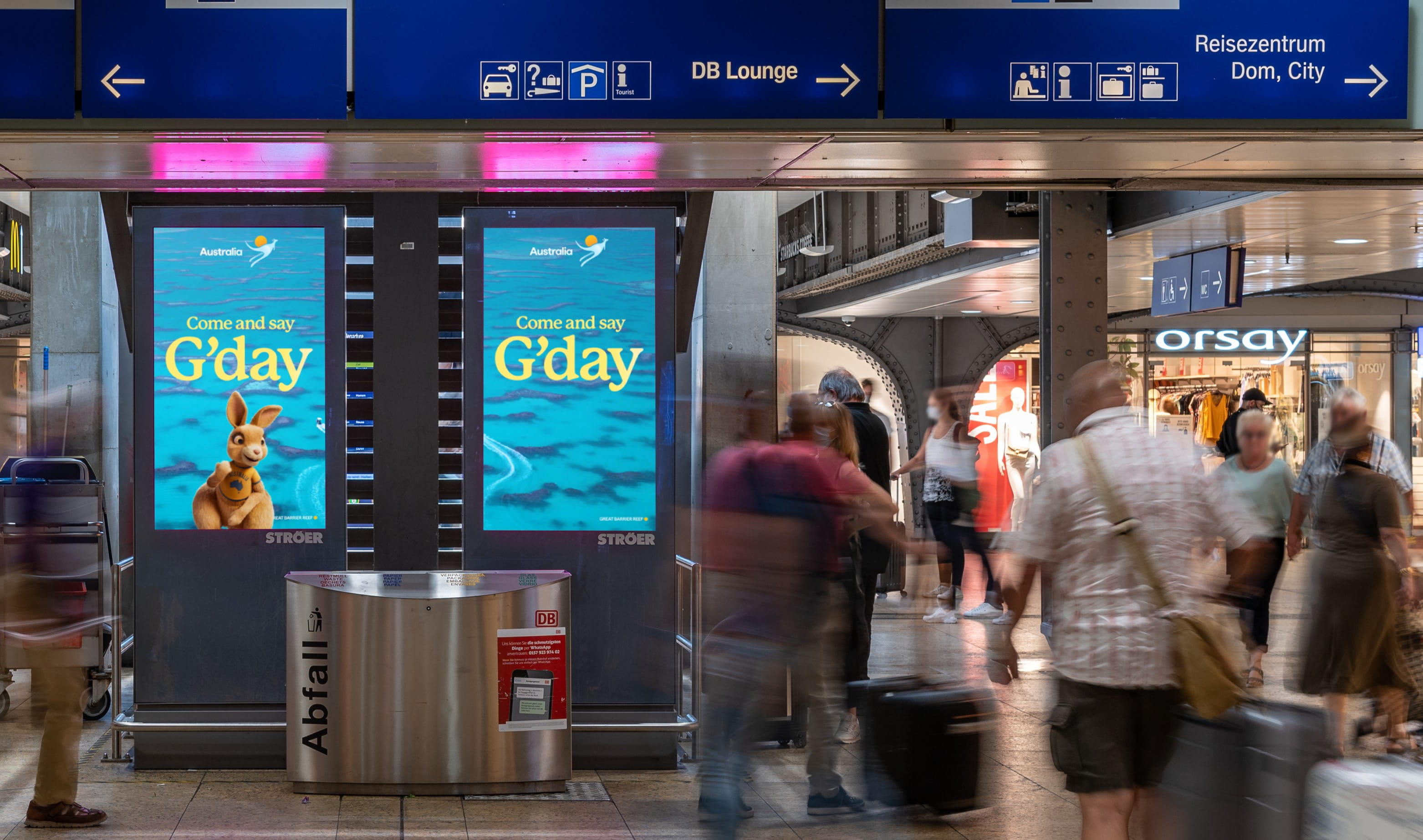 Digitale Außenwerbung von Tourism Australia im Bahnhof mit dem Motiv „Come and say G’day“ und einem Känguru auf DOOH-Screens, vorbeigehende Reisende im Vordergrund.