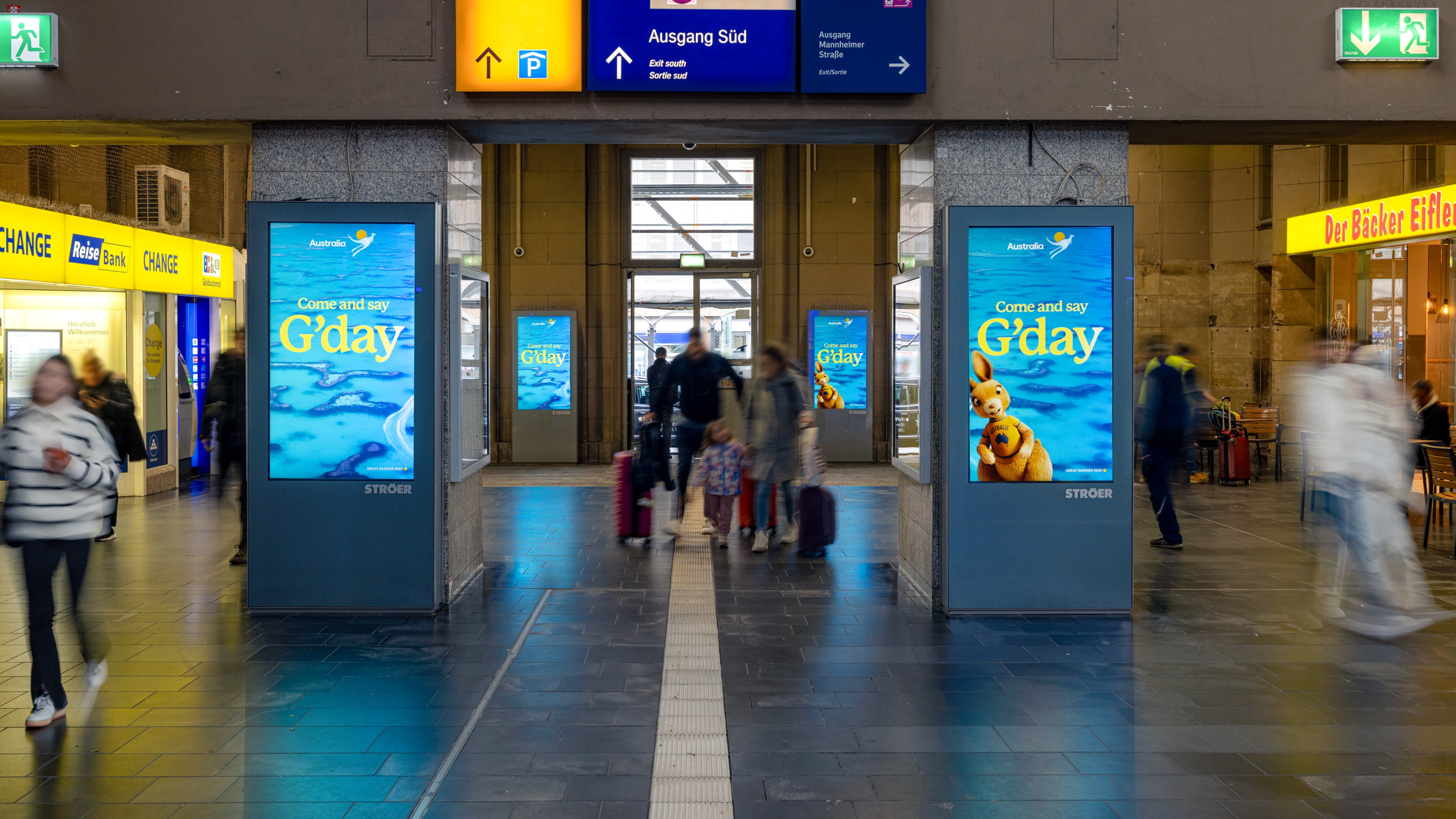 Mehrere digitale Werbescreens von Tourism Australia im Bahnhofsumfeld mit dem Motiv „Come and say G’day“, Reisende mit Gepäck bewegen sich durch die Halle.