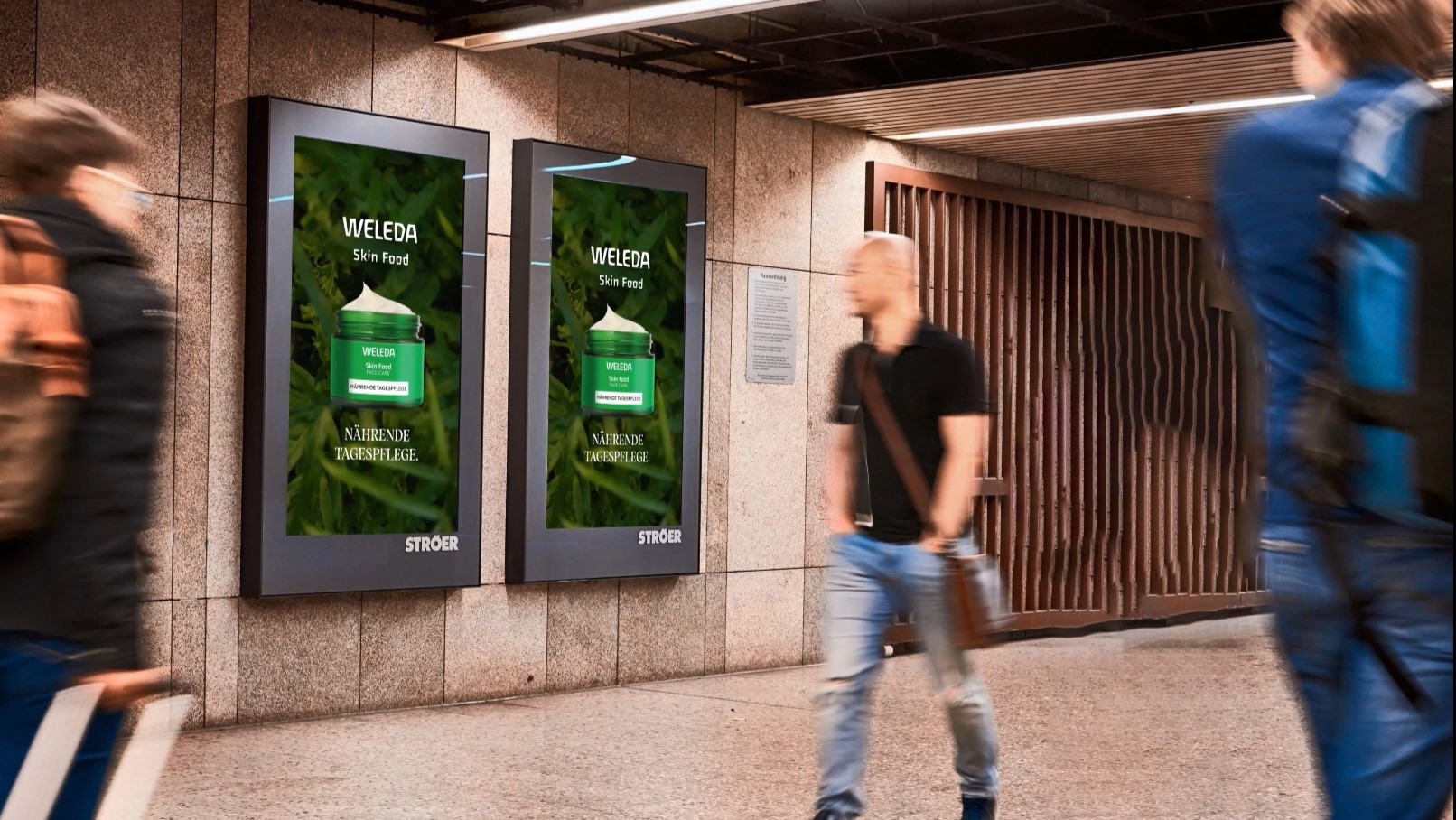 Digitale Displays in Bahnhof mit Weleda Skin Food Werbung – Zwei digitale Werbetafeln präsentieren die Weleda Skin Food Tagespflege mit grünem Naturhintergrund in einer Fußgängerpassage.