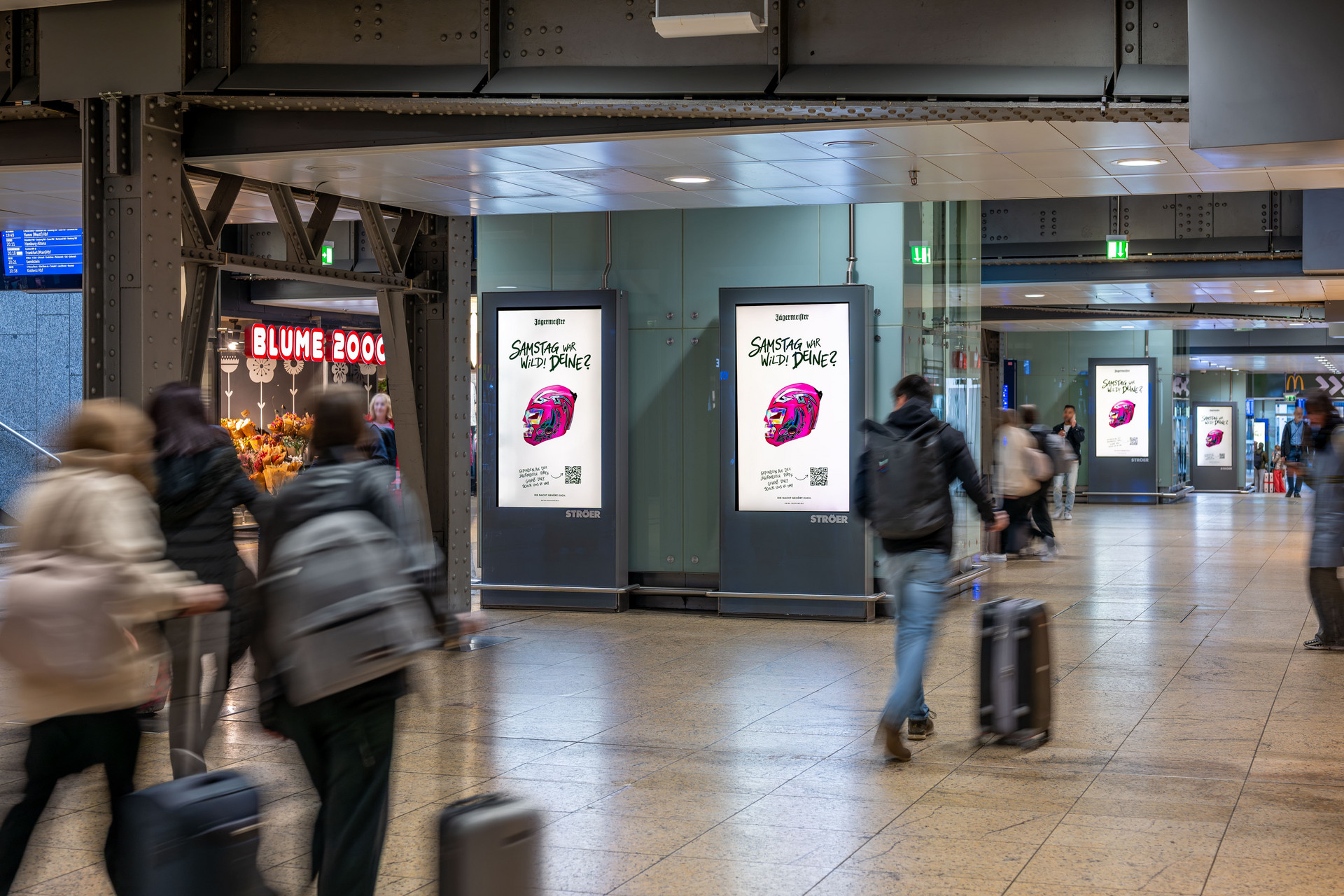 Eine belebte Halle im Kölner Hauptbahnhof mit Reisenden, die mit Koffern durch das Bild laufen und durch die Bewegung verschwommen wirken. Im Zentrum stehen zwei hochkant installierte, digitale Werbescreens nebeneinander. Sie zeigen eine weiße Jägermeister-Werbung mit einem bunten Helm und der Aufschrift „Samstag war wild! Deins?“.